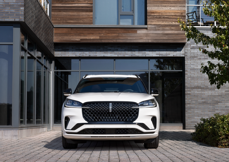 A white 2025 Lincoln Aviator® SUV is shown parked outside a modern building with gray brick and wood accents. | Uptown Lincoln in Wauwatosa WI