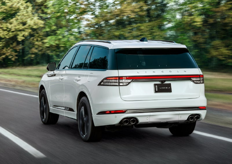 A white 2026 Lincoln Aviator® SUV is being driven away down a road lined with colorful trees. | Uptown Lincoln in Wauwatosa WI