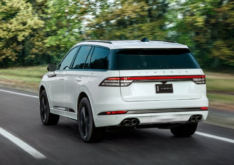 A white 2026 Lincoln Aviator® SUV is being driven away down a road lined with colorful trees. | Uptown Lincoln in Wauwatosa WI