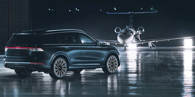 Rear three-quarter view of a black Lincoln Navigator SUV parked on a wet airport hangar floor with a private jet illuminated in the background
