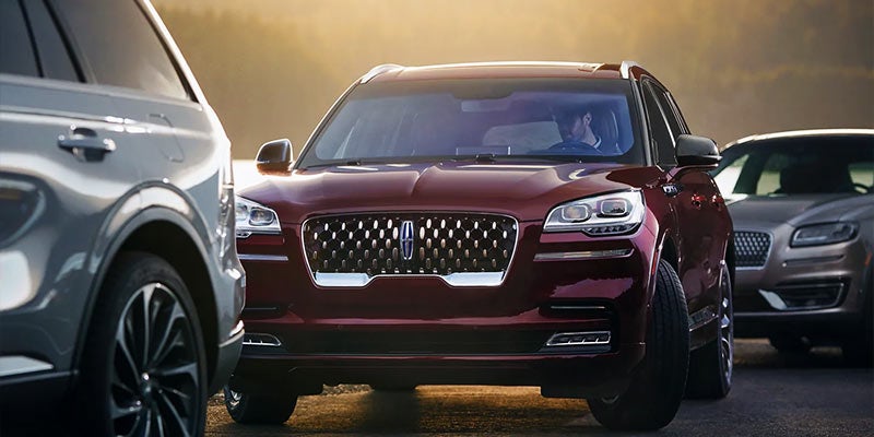 Front view of a burgundy Lincoln Navigator SUV driving between two vehicles on a roadway in warm evening light