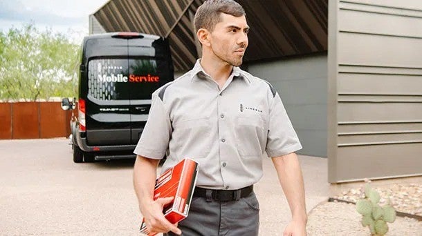 Lincoln service technician in uniform, holding a red binder, walks from a black Mobile Service van towards a building.